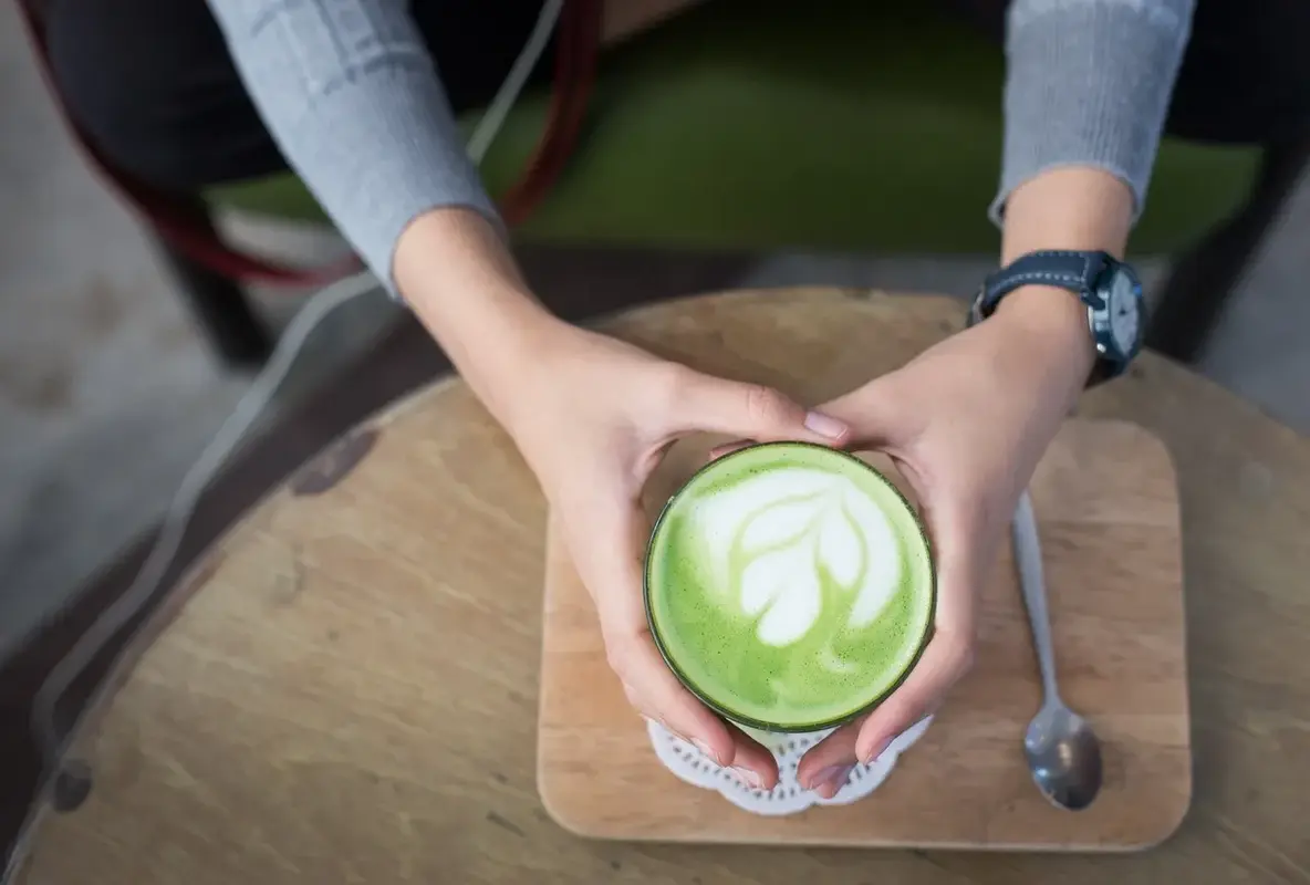 Hands holding a green matcha latte with white leaf design on a wooden table.