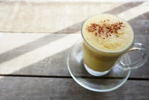 Glass cup of turmeric milk with frothy top sprinkled with cinnamon on wooden table.