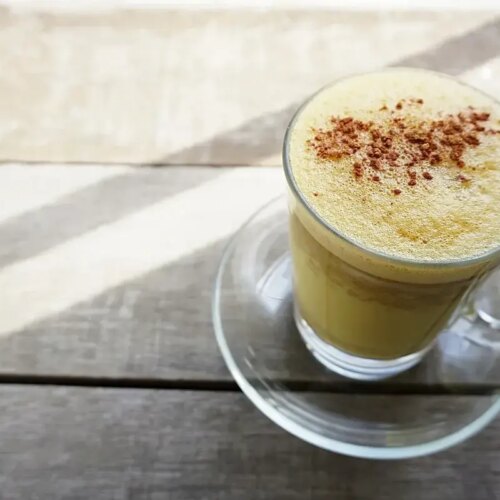 Glass cup of turmeric milk with frothy top sprinkled with cinnamon on wooden table.